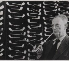 Een man rookt uit een lange pijp van de N.V. Goedewaagen. Op de achtergrond het modellenbord met pijpen, circa 1950