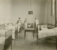 De meest oostelijk gelegen ziekenzaal in het Catharinagasthuis met vrouwelijke patiÃ«nten, artsen en verplegend personeel, plus een kolengestookte kachel, 1910